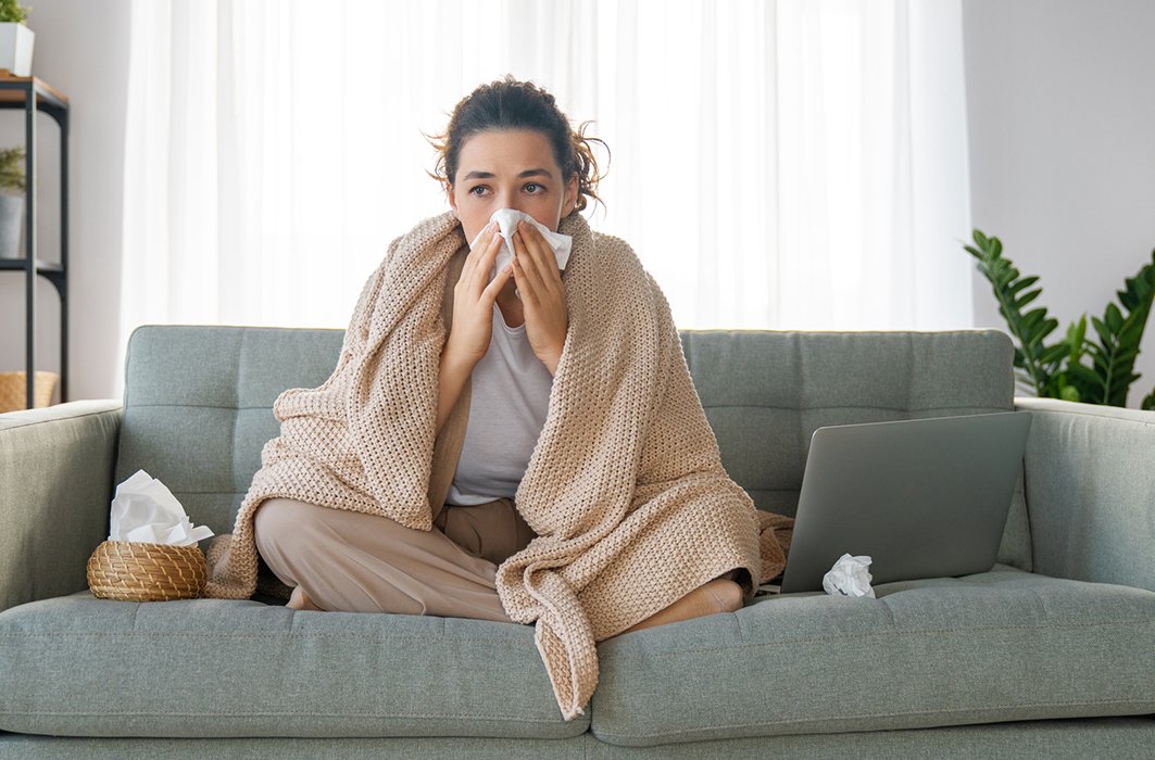 Une personne se mouche, enroulée dans une couverture et assise sur son canapé.
